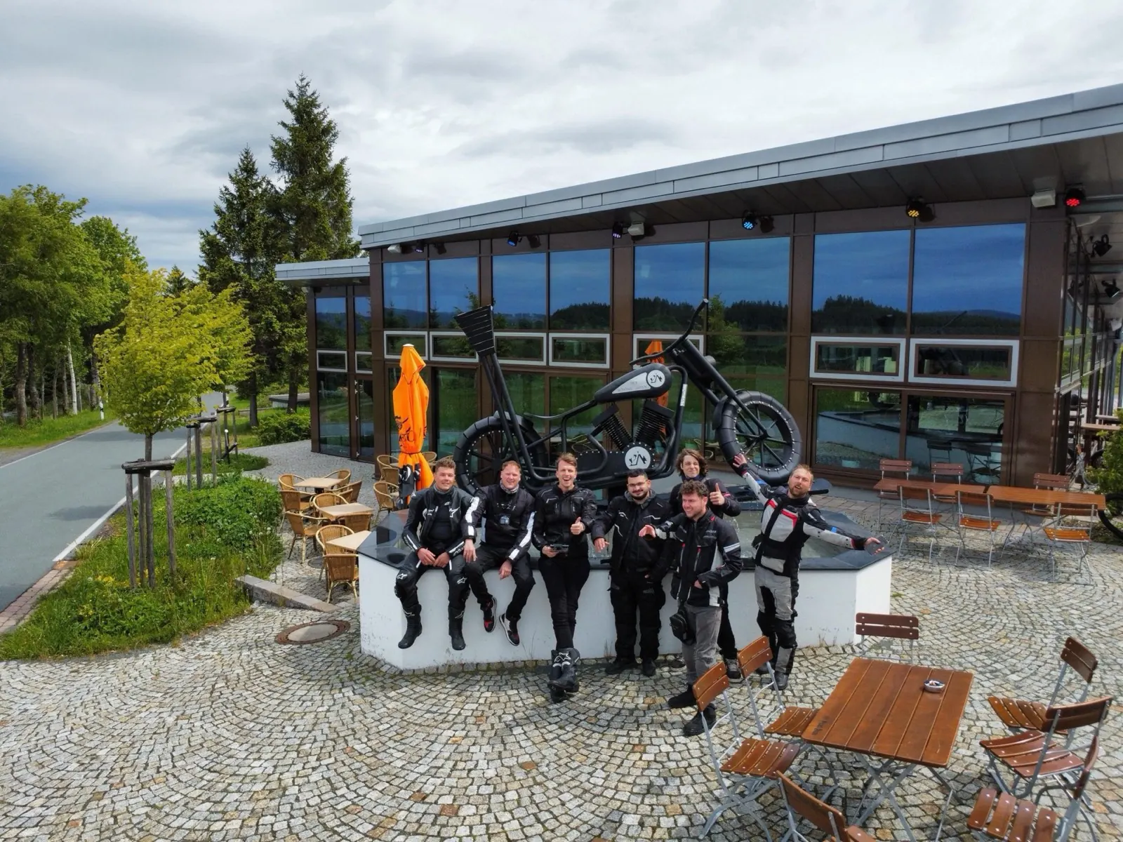 Groep motorrijders poseert op een terras met een groot motorbeeld tijdens een stop onderweg